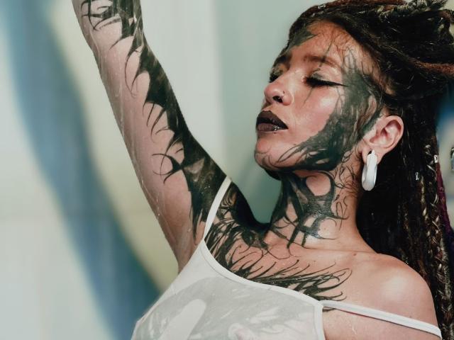 LucyDolly, maquillage foncé, tatouages sur le cou et le bras, débardeur blanc mouillé, bras levé, écouteurs, rouge à lèvres foncé, cheveux tressés, et une expression sexy et intense..