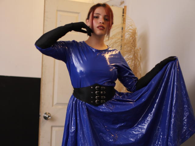 ElizaLovely with red hair wearing a shiny blue latex dress, black corset, and black gloves, holding out the skirt, showing her tits and a confident, sexy look.