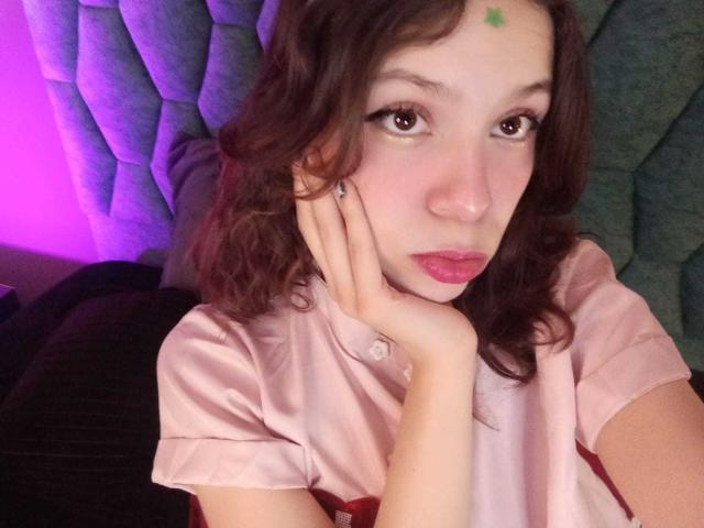 SophieBlake with curly brown hair, pink shirt, green forehead dot, pouty lips, resting hand on cheek, looking up with brown eyes, pink and purple lighting.