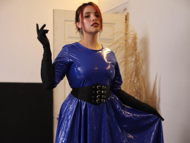 ElizaLovely draagt een glanzende blauwe latexjurk met een zwarte corset en handschoenen, ze houdt haar rok omhoog en laat haar borsten zien, en staat in een kamer met pampa gras..