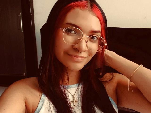 AriaRosel mit roten und schwarzen Haaren, die runde Brillen, einen Nasenring und ein weißes Tanktop trägt, lächelt in die Kamera, hat eine Hand im Haar und trägt ein Armband am Handgelenk..