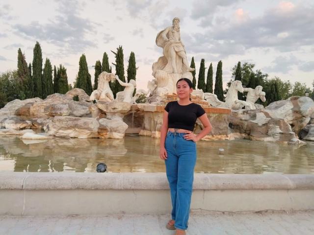 AmaraParker stands in front of a stone fountain, wearing a black top, blue high-waisted jeans, and brown shoes, with a serious expression and hands on her hips.