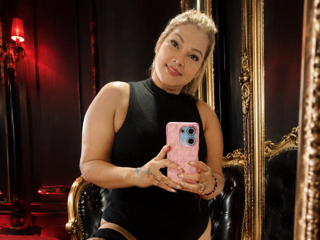 ViancaCastellanos with blonde hair in a black sleeveless top, holding a pink phone, smiling, showing tattoos on her fingers, sitting in a black leather chair.