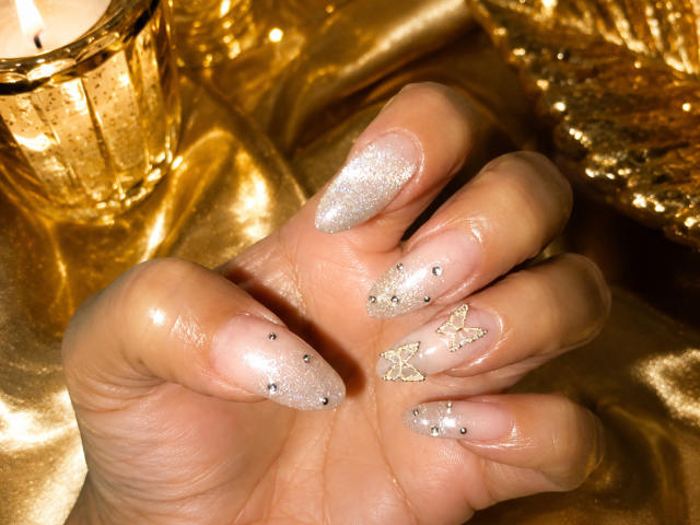 Vianca Castellanos' glänzende, glitzernde, schmeichelnde Nägel mit schimmerndem silbernen Lack, kleinen schwarzen Punkten und goldenen Schmetterlingsmotiven, wobei sie eine Hand über einer goldenen Kerze auf Seide hält..