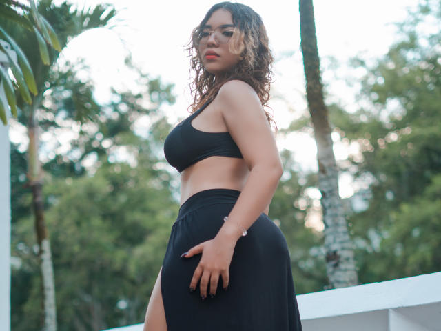 SweetyAliceHotX com cabelo castanho encaracolado, óculos, top de ginástica preto e saia preta, mostrando o bumbum, em pé ao ar livre, com palmeiras ao fundo, olhando por cima do ombro..