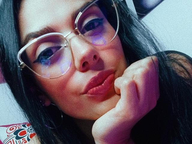 Anaïs Hilton, avec ses lèvres pincées, ses cheveux noirs épais et ses grandes lunettes transparentes, appuyant son menton sur sa main, mettant en valeur son fard à paupières bleu et son rouge à lèvres rose..