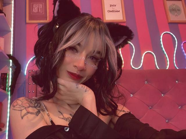 LaurenEvie69 with black cat ears, black off-shoulder top, and tattoos on her shoulder, has red lipstick, white and black hair, and is smiling with her hand on her cheek.