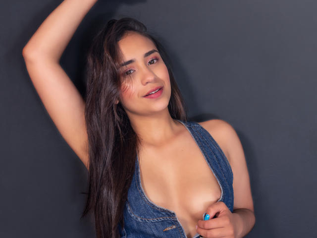 LunaBrittonn with long dark hair, blue eyes, and pink blush, wearing a low-cut denim dress, exposing her tits, one hand above her head, smiling, against a dark background.