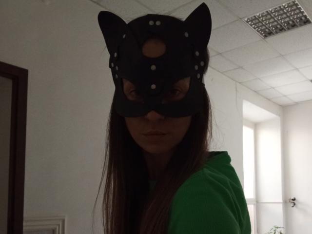 ViktoriiaLu, com cabelos castanhos longos, usando uma máscara preta de gato com orelhas e bolinhas brancas, e um moletom verde, tem uma expressão séria em um quarto com pouca luz..