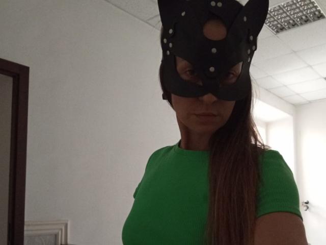 ViktoriiaLu, avec de longs cheveux bruns, portant un masque de chat noir avec des oreilles et des points blancs, et un t-shirt vert côtelé, qui révèle ses seins. Pièce avec des murs blancs..