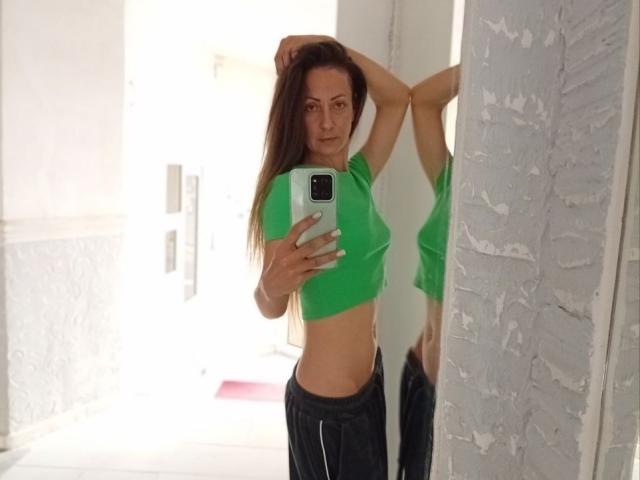 ViktoriiaLu with long brown hair, green crop top, and black pants, standing in front of mirror, arms raised, holding phone, showing her flat stomach and small tits.