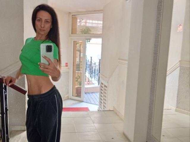 ViktoriiaLu met lang bruin haar, een groene top die haar buikspieren laat zien, zwarte joggingbroek, met een telefoon in haar hand, staand in een gang, een spiegelzelfportret, kijkend naar de camera, met een serieuze uitdrukking..