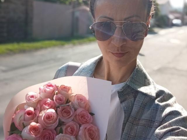 ViktoriiaLu com rosas cor-de-rosa, usando óculos de aviador, camisa xadrez e camiseta branca, segurando um buquê de rosas cor-de-rosa, sorrindo levemente, rua ensolarada ao fundo..