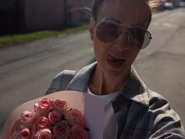 ViktoriiaLu mit rosa Rosen, trägt ein grünes Karohemd und Aviator-Sonnenbrille, hält einen Blumenstrauß, der Mund ist leicht geöffnet, steht auf einer sonnigen Straße..