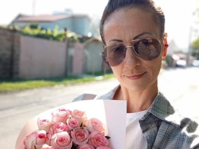 ViktoriiaLu mit rosa Rosen, die eine Sonnenbrille, ein kariertes Hemd und ein weißes Oberteil trägt, hält einen Blumenstrauß in der Sonne, steht auf der Straße und lächelt leicht..