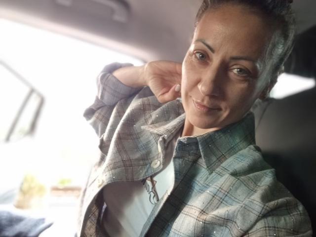 ViktoriiaLu, avec une peau brun clair et des cheveux bruns bouclés, portant une chemise à carreaux, souriant légèrement, la main près de l'oreille, dans une voiture avec la lumière du soleil entrant par la fenêtre..