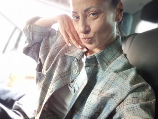 ViktoriiaLu met een nadenkende uitdrukking, gekleed in een geruite blouse, hand op haar wang, zittend in een auto, licht van het raam aan de linkerkant, subtiele glimlach, neusring, donker haar in een knot..