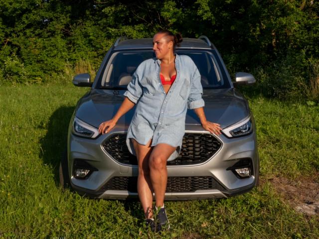 DianaMGG met een bruine huid, bruin haar in een knot, draagt een rode bh, een blauwe jurk met een shirtmodel en sandalen, en zit op de voorgrille van een grijze Nissan SUV..