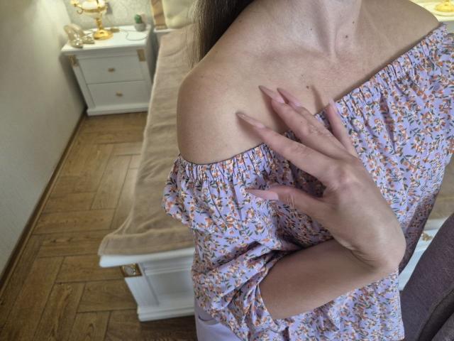 Il vestito floreale con spalline scoperte di Rouzi mette in mostra il suo seno, con una mano che tocca la clavicola. Ha le unghie rosa e un incarnato chiaro. Il letto e il comodino sono sullo sfondo..