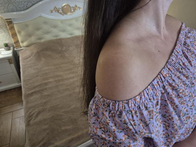 Il vestito floreale con spalline scoperte di Rouzi mette in mostra il suo seno piccolo e la spalla nuda, con lunghi capelli castani, in una camera da letto con un letto beige e un comodino bianco..