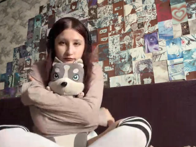 TameikaVannoy with brown hair, wearing headphones, hugging a grey and white stuffed dog, sitting on a dark couch, wearing a beige shirt and white socks with black stripes.