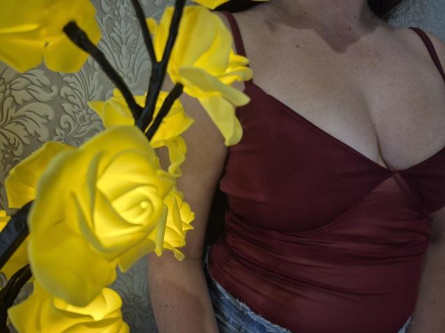 La maglia color borgogna di Rouzi, che mette in risalto il décolleté ed è senza maniche, con i suoi fiori gialli a forma di rosa in primo piano. Il suo seno è ben visibile sotto la maglia. Indossa dei jeans blu..