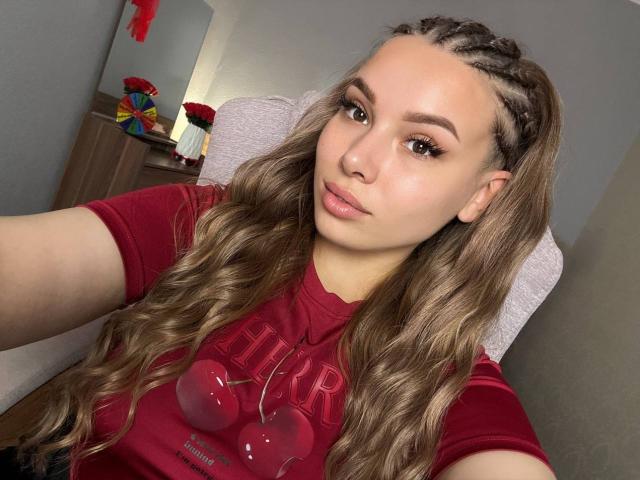 Niferttiti with long, wavy brown hair in braids, wearing a red shirt, pouting lips, brown eyes, and a slight smile, in a selfie. Her ass is not visible.