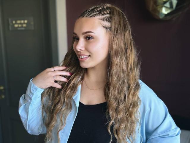 Niferttiti with long wavy brown hair, braided crown, smiling, wearing a light blue shirt over a black top, hand touching her hair, pink nails, subtle makeup.
