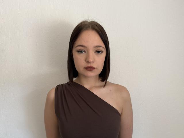 Jestine Baumann, mit schulterlangem, braunem Haar, trägt ein braunes Top mit einem Schulterträger und hat einen neutralen Gesichtsausdruck, eine kleine Nase und dezentes Make-up. Sie steht vor einer weißen, strukturierten Wand..