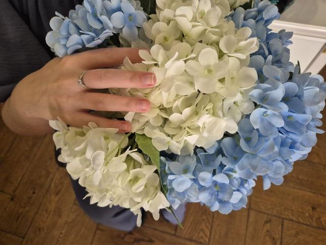 La main de Meeranda, ornée d'une bague en argent, tenant un bouquet d'hortensias bleus et blancs, révélant ses doigts manucurés en rose, sur un plancher en bois..