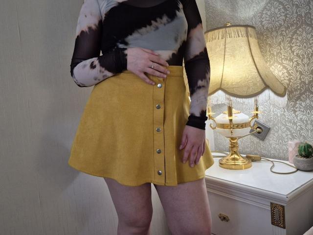 Meeranda lleva una camiseta con estampado de tie-dye, una falda amarilla con botones y tiene una mano sobre su vientre. Mangas negras transparentes, uñas amarillas, una lámpara dorada y una mesita de noche blanca en el fondo..