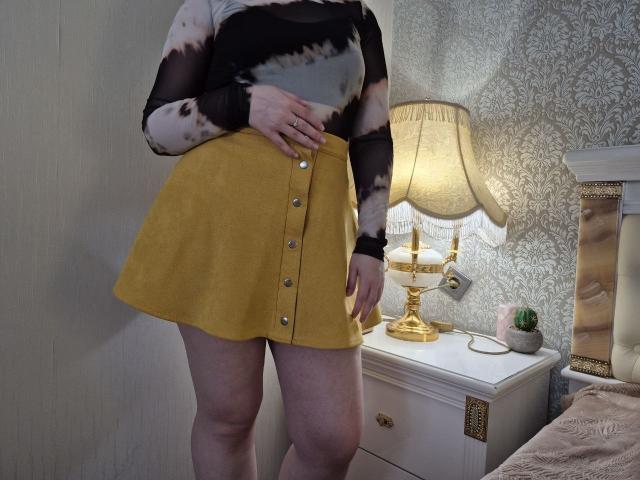 Meeranda wearing a yellow button-up skirt, black sheer long-sleeve top, and no underwear, with her hand on her belly, standing next to a white nightstand with a lamp.