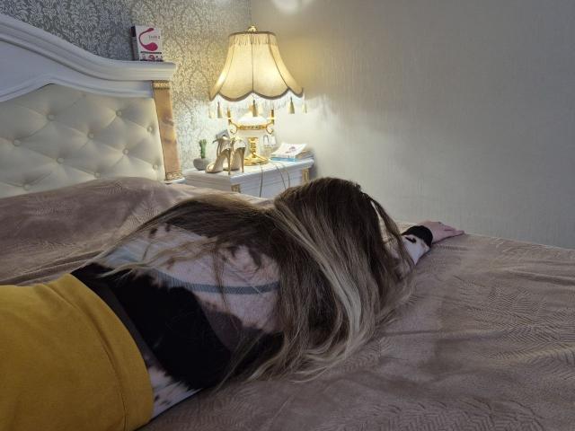 Meeranda, blonde, lying face-down on a bed with a pinkish-brown blanket, wearing a yellow skirt and black-and-white top, with her ass up and head down.