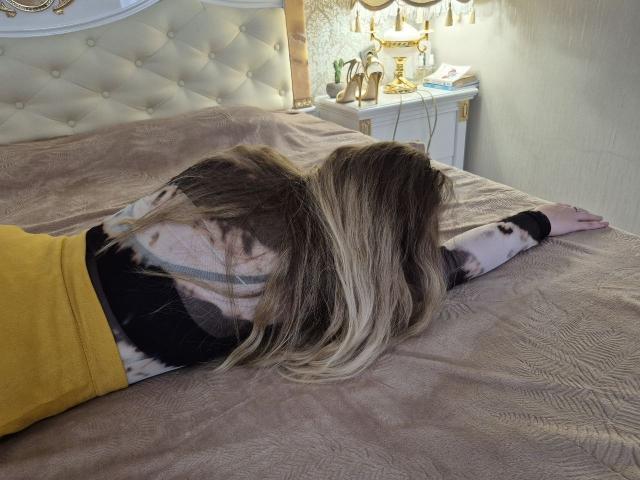 Meeranda's ass is on a bed, face down, arms out, wearing a yellow top, black and white shirt, and blonde hair covering her back.