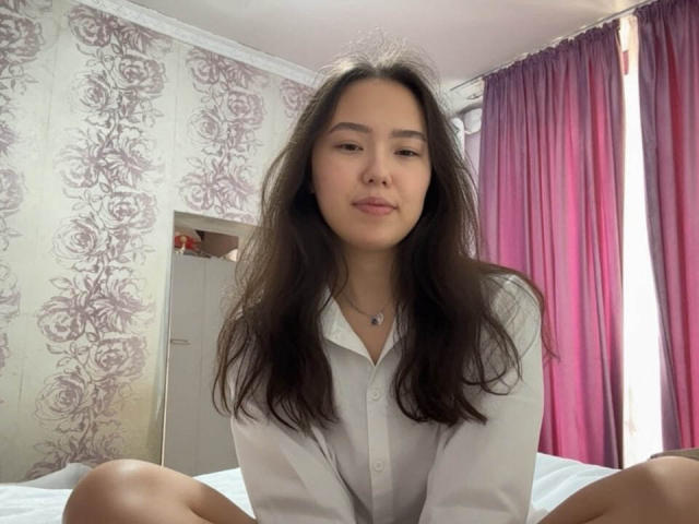 MitsueDorst heeft lang donker haar, een lichte glimlach en draagt een witte blouse met knopen, ze zit op een bed met haar knieën opgetrokken, met paarse gordijnen op de achtergrond..