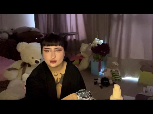 LynnaMitton with black hair, bangs, and bold eyeliner, wearing a black robe, holding a foil-covered object, tattoos visible, surrounded by stuffed animals and a flower arrangement.