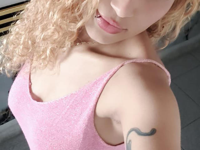 NaomyeLm with curly blonde hair, pink sparkly tank top, and a tattoo on her right arm, showing her pouty pink lips and small tits.