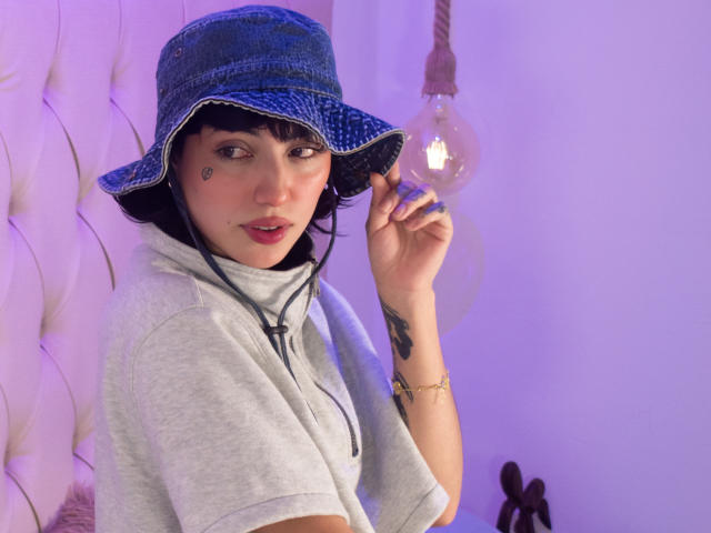 CleoZapata wearing a blue denim bucket hat, gray t-shirt, and headphones, with a light bulb hanging above, has a tattoo on her right arm and a beauty mark.