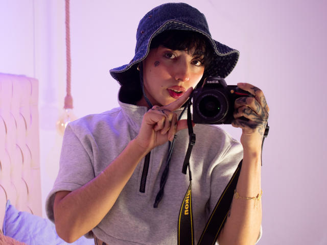 CleoZapata with black hair, wearing a blue bucket hat and gray tee, holding a Canon camera, finger on lip, tattoos on right hand, purple light in background.