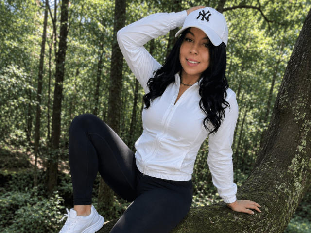 NenaBlack mit langen schwarzen Haaren, einer weißen NY-Kappe, einem weißen Hemd, schwarzen Hosen und weißen Turnschuhen, sitzt auf einem Baum, die Hand in der Kappe, lächelnd, mit einem Wald im Hintergrund, weiße Turnschuhe..