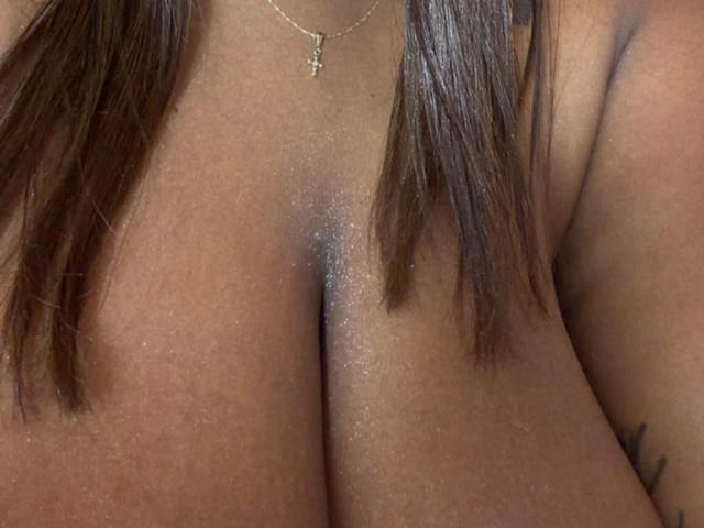 Os seios grandes e brilhantes de NatyLm, com cabelo castanho de ambos os lados, usando um pequeno colar com uma cruz, textura da pele visível e uma tatuagem parcialmente visível no lado direito..