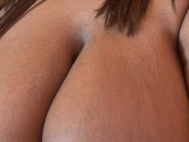 El trasero moreno de NatyLm en un primer plano, mostrando una piel suave y oscura con un pequeño folículo piloso visible cerca de la parte superior. Se puede ver un poco de cabello castaño..