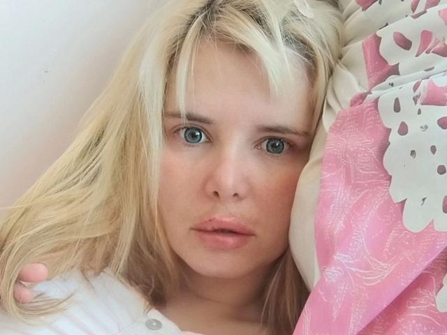 ElenaSchatz-hot a les cheveux blonds, les yeux bleus et les lèvres roses, elle est allongée sur un oreiller rose et blanc à motifs, vêtue d'une chemise blanche, et elle a l'air fatiguée et sérieuse..