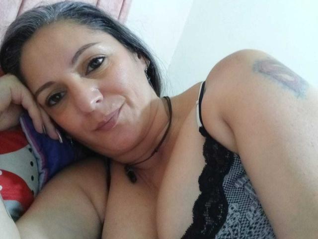 MadameScarlett with dark hair, small nose, and a tattoo on her right shoulder, smiling at the camera, wearing a black lace bra, and a necklace with a dark bead.