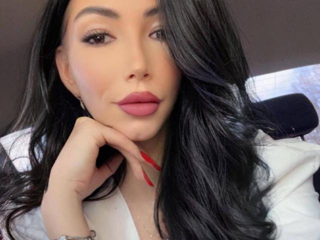 SinReea has long black hair, full pink lips, and red nails, with her hand touching her cheek. She's looking directly at the camera with a slight smile.