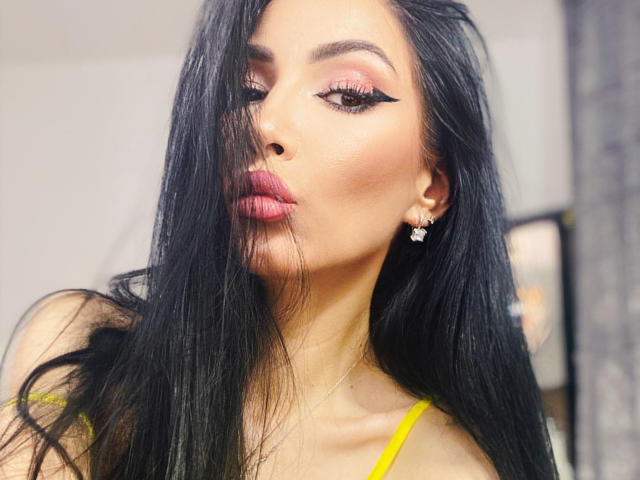 SinReea with long black hair, pink eyeshadow, black winged eyeliner, pink lipstick, yellow top, and diamond earrings, pouting slightly. Her ass is not visible.