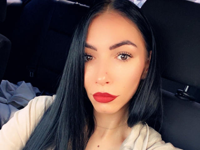 SinReea has long black hair, red lipstick, and brown eyes, wearing a white top, with a nose piercing and a small beauty mark on her chin.