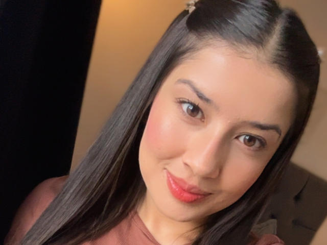 Emma Lombardi a de longs cheveux noirs, des yeux marron et des lèvres rouges avec un léger sourire. Elle porte un haut de couleur rose-brun, et sa peau est lisse avec un teint clair et chaud..