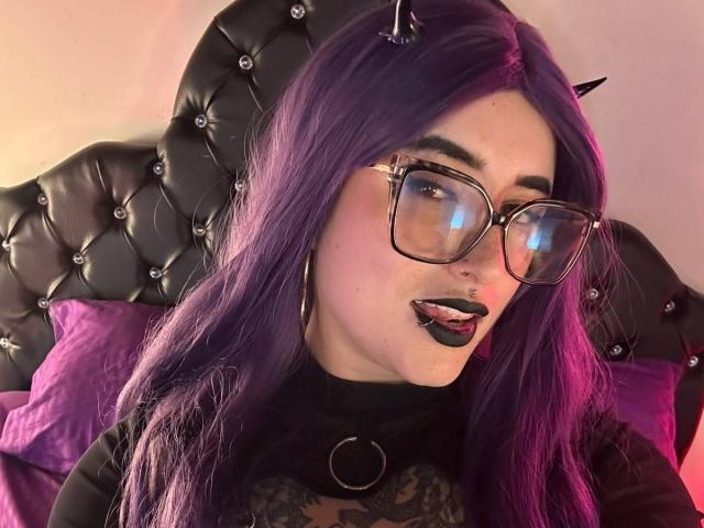 LilithDark69 with purple hair, black horn headband, thick-rimmed glasses, black lipstick, tongue out, nose piercing, black top with ring, sitting on black tufted headboard, purple pillow.
