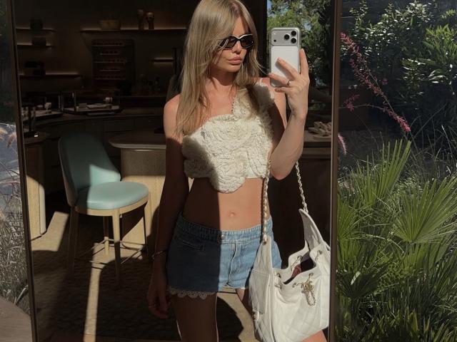 TheHottestBlonde with blonde hair, black sunglasses, white textured crop top, blue denim shorts, white handbag, holding iPhone, standing in sunlight, looking into mirror, showing her flat ass.